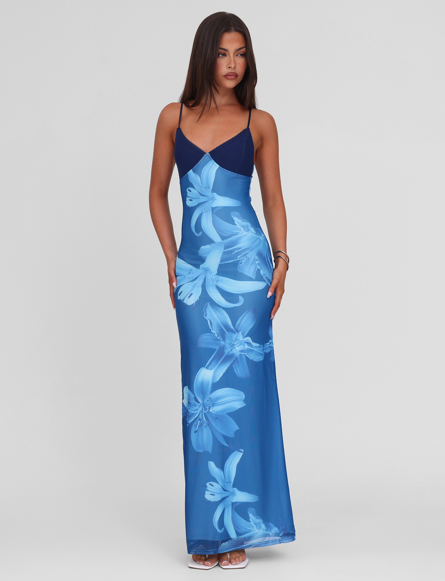 A woman wears the TIGERMIST ALAINA DRESS - BLUE ORCHID, a sleeveless, floor-length maxi with adjustable spaghetti straps and bold light blue floral prints. She pairs it with white sandals and stands before a plain white background, hair flowing.